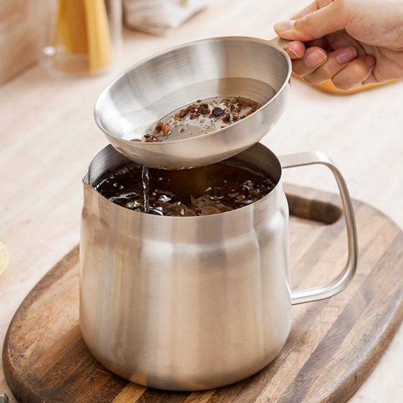 Stainless steel oil pot strainer being used to pour coffee into a mug on a wooden surface.